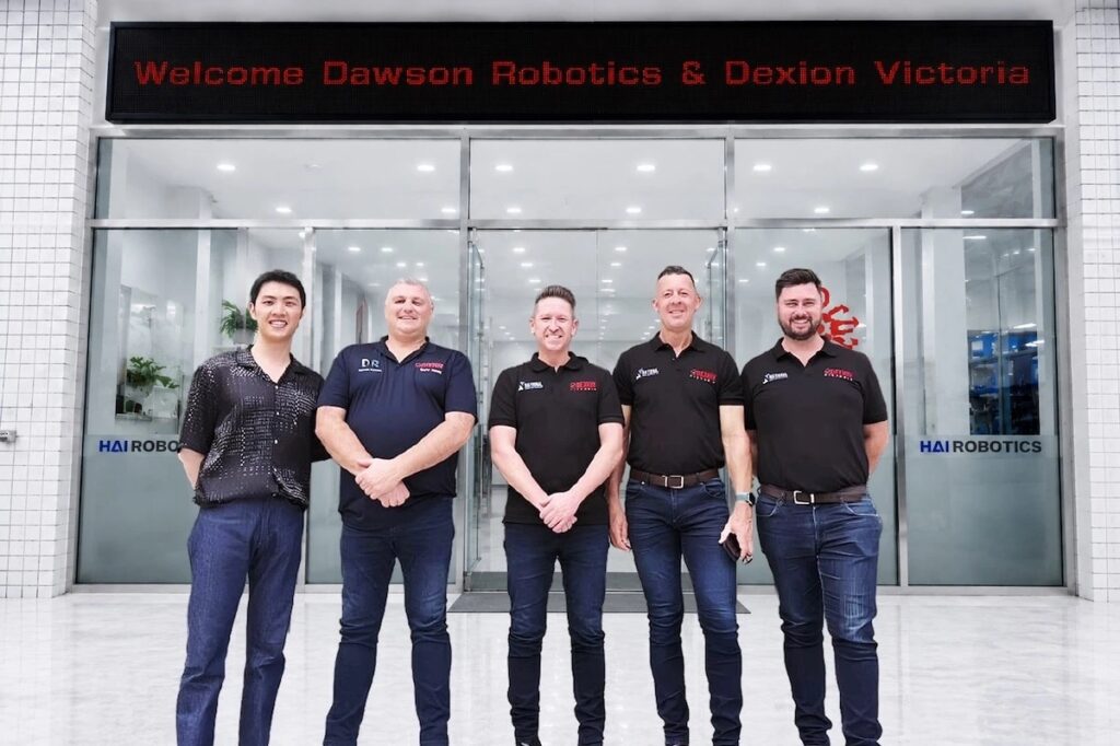 Five people standing at the entrance to Hai Robotics' facility, beneath an illuminated sign reading Welcome Dawson Robotics and Dexion Victoria
